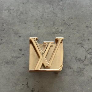 Louis Vuitton LV bältesspänne i guld - Säljer ett exklusivt bältesspänne från Louis Vuitton i guldig metall med den ikoniska LV-loggan. Perfekt för att ge din outfit en lyxig touch. Passar till bälten med utbytbart spänne.