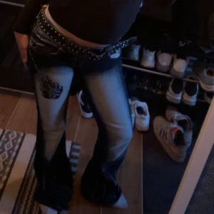 Bootcut jeans med patch och nitar - Snygga bootcut jeans med ljus tvätt och slitningar, dekorerade med en svart patch på låret och nitar i midjan. Byxorna har låg midja och ett brett bälte med silverdetaljer för extra edge.