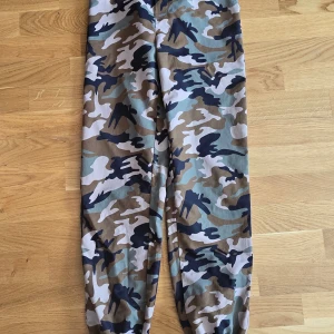 Cargobyxor med camouflage-mönster - Säljer ett par cargobyxor med klassiskt camouflage-mönster i grönt, beige, svart och grått. Byxorna har resår vid bensluten och är perfekta för en avslappnad streetstil.