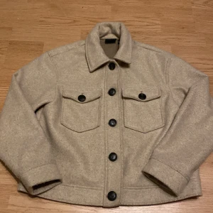 Beige overshirt från Vero Moda - Säljer en beige overshirt från Vero Moda med klassisk krage och fyra stora svarta knappar framtill. Jackan har två bröstfickor med lock och knappar samt långa ärmar. Perfekt för lager-på-lager-stil under höst och vår.