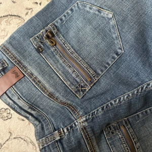 Blå bootcut jeans med dragkedjedetaljer - Säljer ett par blå jeans med bootcut passform och snygga dragkedjor på fickorna både fram och bak. Jeansen har klassisk femficksdesign och bruna sömmar som kontrast. Perfekta för dig som gillar en lite retro vibe och vill sticka ut med detaljer. Längd 34 passar en storlek 38 i midjan! 