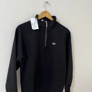 Svart halvzip tröja från Lacoste - Svart tröja från Lacoste med halv dragkedja och klassisk broderad krokodil på bröstet. Tröjan har hög krage och är tillverkad i mjukt bomullsmaterial. Perfekt för en stilren och sportig look. Fick den som present och jag hade en exakt samma