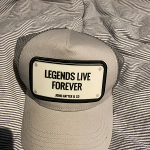 Grå keps från John Hatter & Co - Snygg grå keps med svart och vit patch framtill med texten 'Legends Live Forever'. Kepsen har mesh baktill och justerbar knäppning för perfekt passform. Perfekt accessoar för en cool och avslappnad stil. Använd ungefär 3 gånger och väldigt bra skick,hör av er vid funderingar.pris kan diskuteras 