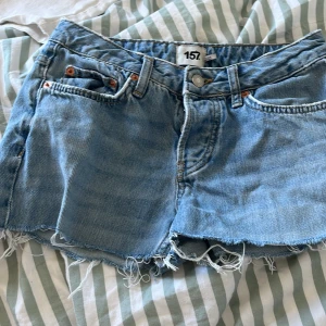 Ljusblå jeansshorts med fransar från 157 - Säljer ett par ljusblå jeansshorts från 157 med råa fransade kanter och klassisk femficksmodell. Shortsen har knapp- och dragkedjestängning samt bälteshällor. Perfekta för varma dagar!