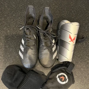 Svarta fotbollsskor från Adidas med benskydd och strumpor - Säljer ett par svarta Adidas fotbollsskor med vita detaljer och dobbar, tillsammans med benskydd och svarta strumpor. Skorna har snörning och är perfekta för fotbollsträning eller match. Skorna är i storlek 37,5 och är i bra skick. Strumporna är aldrig använda.