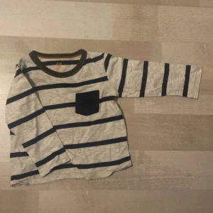 Randig långärmad tröja med bröstficka - Grå långärmad tröja från H&M med mörkblå horisontella ränder och en matchande mörkblå bröstficka. Tröjan har rund halsringning med mörk kant och är tillverkad i mjuk bomull. Barn tröja storlek 92