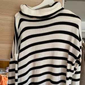 Randig stickad polotröja - Säljer en vit och svart randig stickad polotröja med lång ärm och hög krage. Perfekt för att hålla sig varm och snygg under kyliga dagar. Klassisk och enkel design som passar till mycket.