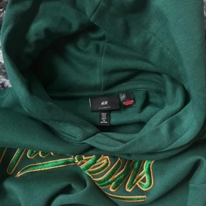 Grön Stranger Things hoodie H&M (M) - Snygg grön hoodie från H&M x Stranger Things med broderad 'Hawkins' text i gult och grönt på bröstet och 'Stranger Things' broderat på ärmen. Tröjan har huva, känguruficka och relaxed fit. Perfekt för dig som älskar serien och vill ha en chill stil. Köpt för runt 400+ när den kom ut. Aldrig använd 