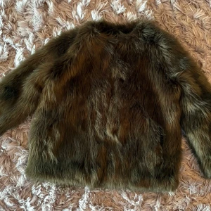 Fuskpälsjacka från By Malina - Säljer en fluffig  pälsjacka från By Malina. Jackan har lång ärm, rund halsringning och är helfodrad. Perfekt för dig som vill sticka ut med en lyxig och trendig look. Färgen är djupgrön brun och materialet känns mjukt och varmt.