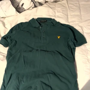 Grön Lyle & Scott piké t-shirt - Snygg grön piké från Lyle & Scott i storlek S. Klassisk krage, tre knappar och den ikoniska gula fågeln broderad på bröstet. Perfekt för dig som gillar stilrena och sportiga plagg. T-shirten är gjord i mjuk bomull och har korta ärmar.