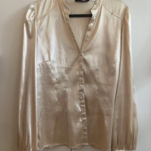 Champagnefärgad silkesblus med knappar - Snygg champagnefärgad blus i silkesliknande material med v-ringning och långa ärmar. Blusen har knappar framtill och vid ärmsluten, samt lätt puffade axlar för en elegant touch. 