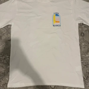 Vit t-shirt från Casablanca M - Casablanca t-shirt i vitt med färgglad tryckt logga på bröstet. Klassisk rund hals och korta ärmar. Materialet är mjuk bomull och trycket har inslag av blått, gult och orange. Perfekt för dig som gillar clean stil med en twist.