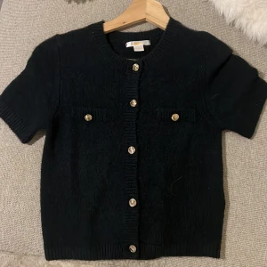 Marinblå kofta med guldknappar Gina Tricot - Kortärmad svart kofta från Gina Tricot med snygga guldknappar framtill och dekorativa fickor. Koftan är stickad och har ribbade kanter vid ärmslut och hals. Perfekt för dig som vill ha en stilren och trendig look. Har ingen prislapp kvar men aldrig använd. 