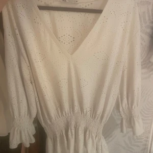 Vit broderad tröja från Zara - Superfin vit tröja från Zara med broderade detaljer och v-ringning. Klänningen har trekvartsärmar med resår och volang, samt smock i midjan för en snygg passform. Perfekt för sommarens alla tillfällen!