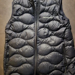 Svart dunväst Peak Performance 170 - Svart dunväst från Peak Performance med vågformat quiltat mönster. Västen har dragkedja framtill, två sidofickor med dragkedja och diskret logga på bröstet. Perfekt för lager-på-lager och outdoor-vibe.