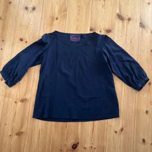 Svart blus från Greta - Enkel och stilren mörkblå blus från Greta med rund halsringning och trekvartsärmar. Blusen har en rak passform och är tillverkad i ett mjukt tyg som ger en skön känsla. Perfekt att matcha med jeans eller kjol för en clean look. Saknar en knapp på ärmen men inget man tänker på.