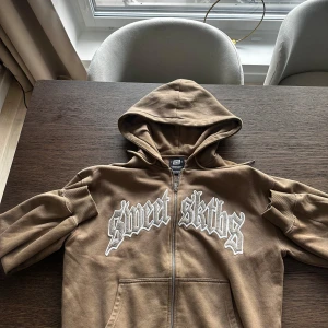 Brun hoodie med dragkedja - Säljer en brun hoodie från Sweet Sktbs med vit broderad text på bröstet. Tröjan har huva, dragkedja framtill och två fickor. Tillverkad i mjuk bomull och har långa ärmar med muddar. Perfekt för en chill och avslappnad stil.
