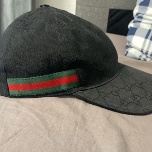 Svart Gucci keps  - Snygg svart keps från Gucci med diskret GG-logomönster över hela kepsen. På sidan finns en klassisk grön och röd rand som ger extra lyxig känsla. Kepsen har böjd skärm och justerbar rem baktill. Perfekt för dig som vill ha en exklusiv streetstil. Har använts i 2 månader. 