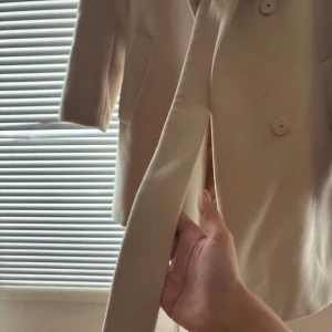 Beige kappa med fuskpäls från H&M - Säljer en beige kappa från H&M i storlek 38. Jackan har en bred krage i fuskpäls, knappar framtill, två fickor och ett bälte i midjan. Perfekt för dig som vill ha en stilren och mysig look under kallare dagar.