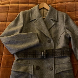 Grå ullkappa från Tiger of Sweden - Säljer en stilren grå kappa från Tiger of Sweden i ull med klassisk krage, dubbelknäppning och bälte i midjan. Kappan har stora fickor framtill och ett bälte med rutigt mönster som ger extra edge. Perfekt för kyliga dagar!