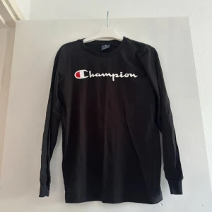 Svart långärmad Champion tröja - Svart långärmad tröja från Champion med stort vitt och rött logotryck över bröstet. Klassisk rund halsringning och mjukt bomullsmaterial som är skönt mot huden. Perfekt för dig som gillar sportig och enkel stil.