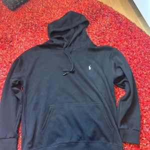 Svart hoodie från Polo Ralph Lauren - Svart hoodie från Polo Ralph Lauren med klassisk logga broderad på bröstet. Tröjan har huva med snörning, stor magficka och långa ärmar. Mjuk bomullskvalitet som är skön att ha på sig. Perfekt för chill eller när du vill ha en clean look.