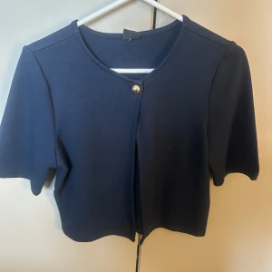 Mörkblå kofta från Gina Tricot XS - Kort, mörkblå kofta från Gina Tricot med en snygg guldig knapp framtill. Modellen har rund halsringning och korta ärmar, perfekt för lager-på-lager. Enkel och stilren design som passar till många outfits.