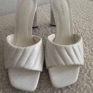 Nya. Vita klackade sandaler från Bershka - Snygga vita sandaler från Bershka med bred, quiltad rem och fyrkantig tå. Skorna har hög blockklack och är tillverkade i syntetmaterial. Perfekta för dig som vill sticka ut med en trendig och stilren look. Storlek 35 men är stora i storlek, är som 36, innelängd 23cm.