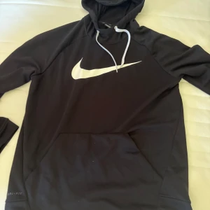 Svart Nike hoodie med vit logga - Svart hoodie från Nike med stor vit Swoosh-logga på bröstet. Tröjan har huva med vita snören och en stor magficka. Tillverkad i Dri-FIT-material som andas, perfekt för chill eller träning. Långärmad och klassisk passform.