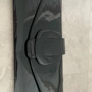 Svart lackad kuvertväska - Snygg svart kuvertväska i glansigt lackmaterial med stilren design och dekorativt lock. Perfekt storlek för mobilen och det viktigaste. Väskan har en elegant form och stängs med ett lock som har en bred detalj framtill.