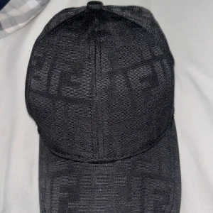 Svart Fendi keps  - Snygg svart keps från Fendi med diskret ton-i-ton logomönster över hela kepsen. Klassisk böjd skärm och justerbar metallspänne bak. Materialet är mjukt och har en lyxig känsla, perfekt för dig som gillar designeraccessoarer med stil.