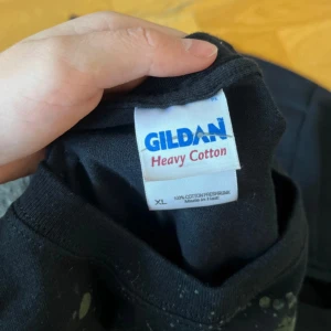 Metallica Stockholm t-shirt XL Gildan - Svart Metallica t-shirt från Gildan i storlek XL, med stort tryck i gult och grått på både fram- och baksidan. Motivet har splattereffekt och text om Stockholm, samt Metallica-logga. Tillverkad i 100% bomull, klassisk passform och korta ärmar.