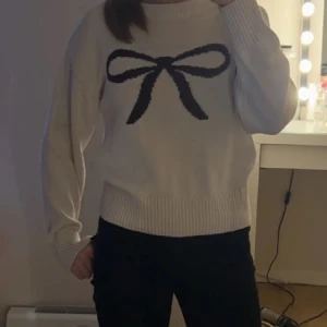 Vit stickad tröja med rosett - LÅNAD BILD! Säljer en söt vit stickad tröja med en stor mörk rosett på bröstet. Tröjan har rund hals, ribbade muddar och långärmad passform. Perfekt för dig som gillar gulliga detaljer och vill ha en mysig look. Postar alltid samma dag som köp 🩷Pris kan diskuteras 💐 storlek 170