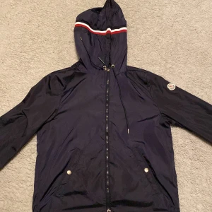 Mörkblå vindjacka från Moncler med huva - Snygg svart vindjacka från Moncler med huva och dragkedja. Jackan har en diskret logga på ärmen, elastiska muddar och en ficka framtill med knapp. Huvan har en cool detalj med röd och vit rand. Perfekt för blåsiga dagar. Nfc scannen funkar också.  Storlek är 2 som motsvarar S och M