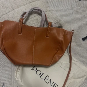 Poléne inspirerad väska - Snygg handväska i brunt/orange skinn med stilren design och guldfärgad logga. Väskan har både handtag och axelrem, samt en praktisk innerficka med dragkedja. Helt ny!🩷