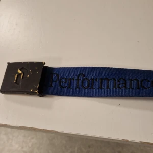 Blå Peak Performance bälte - Snyggt blått bälte från Peak Performance med svart metallspänne och tydlig logga längs bandet. Bältet är gjort i slitstark polyester och har en enkel, sportig design som passar perfekt till jeans eller chinos.