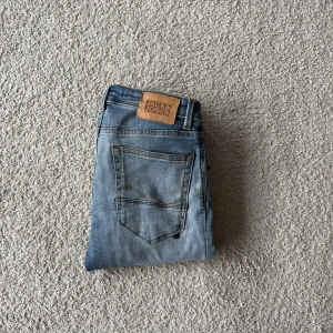 Ljusblå slim  jeans Jack & Jones  - Snygga ljusblå skinny jeans från Jack & Jones i storlek 158. Jeansen har slitningar och ett hål på ena knät för en cool, avslappnad look. Klassisk femficksmodell med dragkedja och knapp. Perfekta till sneakers och hoodie.