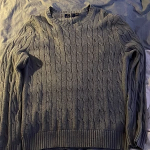 Grå kabelstickad tröja Polo Ralph Lauren (pris kan diskuteras) - Kabelstickad grå tröja från Polo Ralph Lauren i klassisk modell. Tröjan har rund halsringning, ribbade muddar och diskret broderad logga på bröstet. Perfekt för lager-på-lager och chill höst- eller vinterstil.