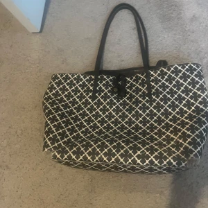 By Malene Birger - Stor shoppingväska i svart och vitt med grafiskt mönster och svarta handtag. Väskan har en rymlig form och stängs med ett spänne upptill. Perfekt för dig som gillar att bära med dig mycket och vill sticka ut med ett coolt print.
