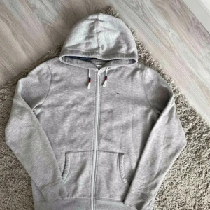 Grå hoodie från Tommy Hilfiger Denim XL - Snygg ljusgrå hoodie från Tommy Hilfiger Denim i storlek XL. Tröjan har dragkedja, huva med dragsko i vitt med röda och marinblå detaljer samt klassisk logga broderad på bröstet. Perfekt för chill dagar och enkel att matcha med jeans eller joggers.