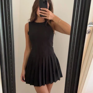 Svart ärmlös plisserad kortklänning - Supersnygg svart kortklänning med plisserad kjol och ärmlös design. Klänningen har en rund halsringning och sitter tight upptill med en loose kjol. Perfekt för dig som gillar stilrena och trendiga plagg med lite edge.