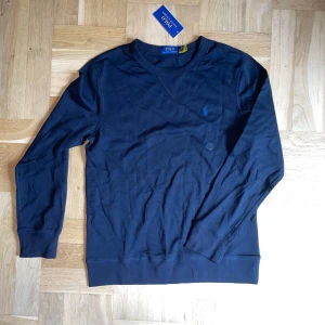 Mörkblå tröja från Polo Ralph Lauren - Snygg mörkblå långärmad tröja från Polo Ralph Lauren i mjuk bomull. Klassisk crewneck med broderad blå logga på bröstet. Perfekt basic-plagg med stilren look och bekväm passform.