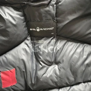 Svart dunjacka från Sail Racing - Svart dunjacka från Sail Racing med huva och horisontella sömmar. Jackan har dragkedja framtill och två fickor med dragkedja. Materialet är glansigt och jackan har en clean look med diskret logga på bröstet. Perfekt för kalla dagar när du vill hålla stilen. Passar alltifrån 160-165, skicket är hållbart men den har hål, funkar perfekt.