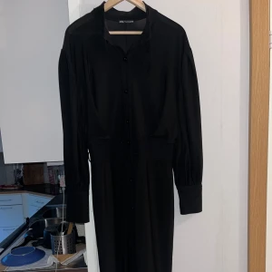 Svart långklänning från Zara - Snygg svart långklänning från Zara med knappar hela vägen framtill och markerad midja. Klänningen har långa ärmar med breda manschetter och klassisk krage. Perfekt för dig som gillar stilrena och tidlösa plagg.