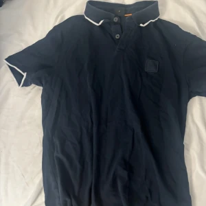 Mörkblå pikétröja från Boss - Snygg mörkblå pikétröja från Boss med vit kant på krage och ärmslut. Klassisk design med korta ärmar, två knappar vid halsen och diskret Boss-logga på bröstet. Perfekt för en chill och stilren look.