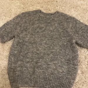 Grå stickad tröja från Inatricot - Mysig grå stickad tröja från ginatricot i storlek XS. Tröjan har korta ärmar och en klassisk rund halsringning. Perfekt för lager-på-lager och chill dagar. Den stickade strukturen ger en skön och avslappnad vibe.