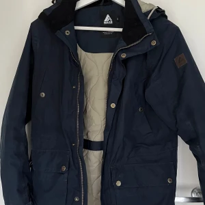 Marinblå Höst och Vinterjacka - Säljer denna marinblåa jackan med beige innetyg som passar perfekt inför det kallare vädret. Den är väl använd men fortfarande i gott skick och har inga tecken på slitage. Går att justera passformen i midja och jackans nedre kant med hjälp dragskor. Jacka  knäppa med dragkedja och knappar och den har fyra fickor på framsidan. Storleken är 160.