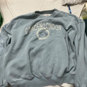 Blå sweatshirt Camden London Pull&Bear - Blå sweatshirt från Pull&Bear med tryck 'Camden London' och grafiskt emblem på bröstet. Tröjan har rund hals, långa ärmar och ribbade muddar. Perfekt för en chill och avslappnad stil.