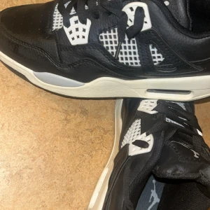 Nike Air Jordan 4 svart/vit sneakers - Snygga Nike Air Jordan 4 sneakers i svart och vitt med klassisk Jumpman-logga på hälen. Skorna har svart skinn, vita meshdetaljer och svarta snören. Den ikoniska siluetten och den tjocka sulan ger en sportig vibe. Perfekta för dig som vill sticka ut med stil.