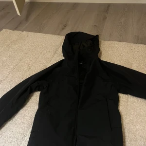Svart vindjacka med huva - Säljer en svart vindjacka med huva och dragkedja framtill. Jackan har en stilren look och är perfekt för blåsiga dagar. Långärmad modell med enkel design och praktiska fickor på sidorna. Passar dig som gillar minimalistisk stil.